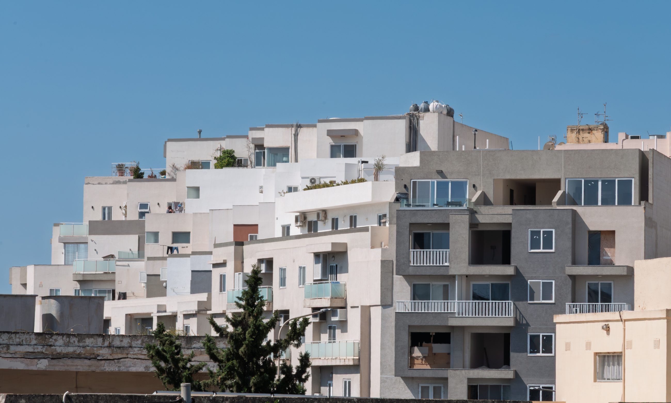 Sunlit white and grey apartment blocks stepping up a hillside under a clear blue sky